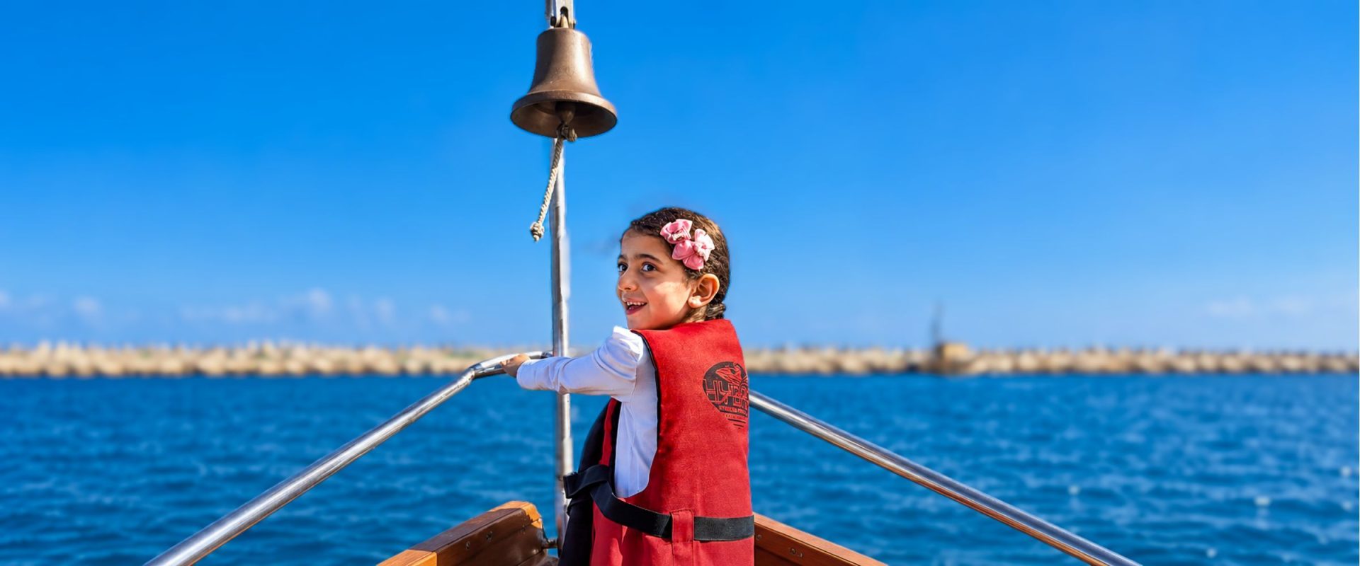 A girl in a red life jacket stands at the bow of a boat and looks back smiling, with the blue sea and a clear sky behind her.